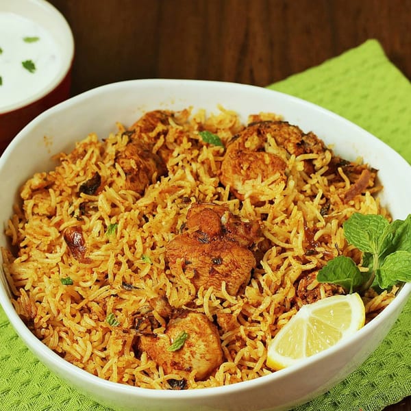 Chicken biryani with rice, chicken pieces, mint, and a lemon wedge, served with raita.