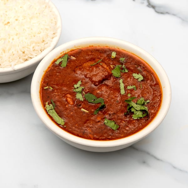 A bowl of rich red Indian curry with cilantro, next to a bowl of white rice.