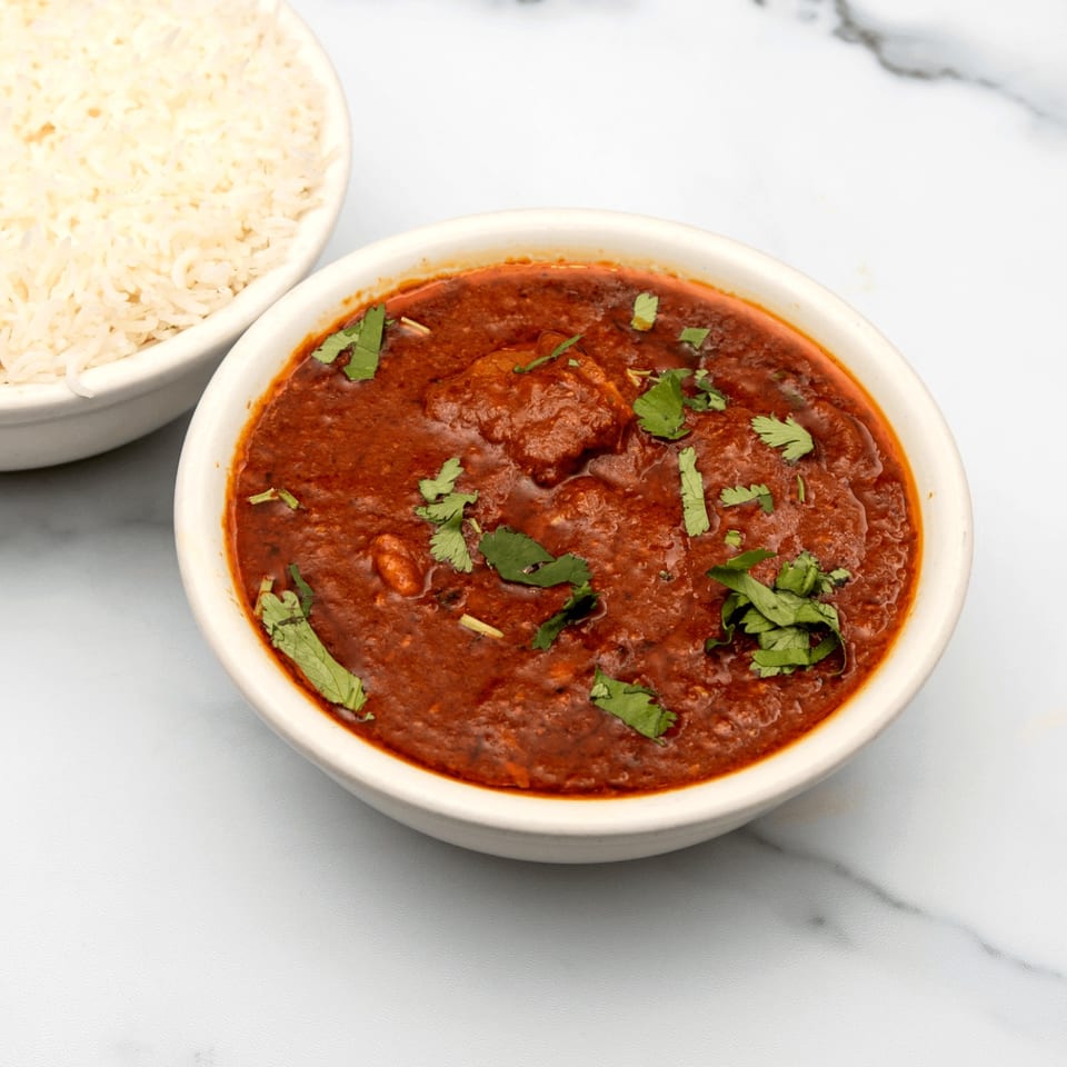 A bowl of rich red Indian curry with cilantro, next to a bowl of white rice.