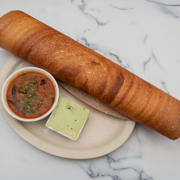 A long, rolled dosa on a plate with a bowl of red sambar and a small dish of green chutney.