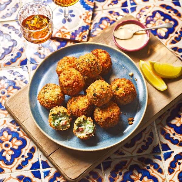 Crispy golden croquettes on a blue plate with lemon wedges, sauce, and drinks on a tiled table.