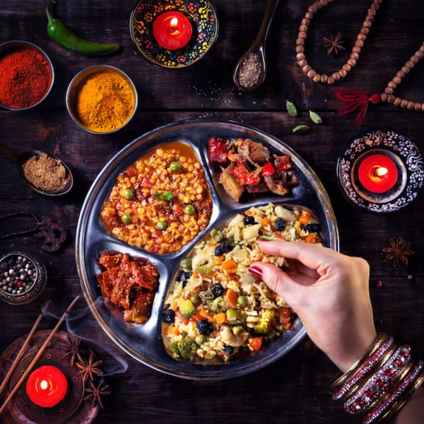 A hand with bangles eats from a thali plate with various Indian dishes and spices.