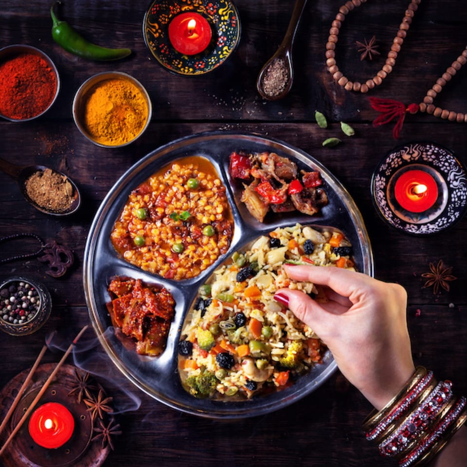 A hand with bangles eats from a thali plate with various Indian dishes and spices.