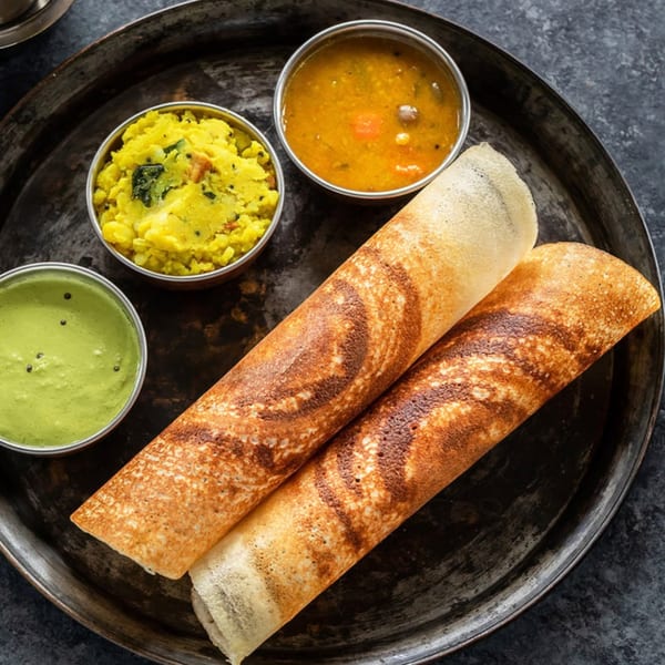 Two golden-brown dosas with bowls of green chutney, yellow potato curry, and sambar on a dark metal tray.