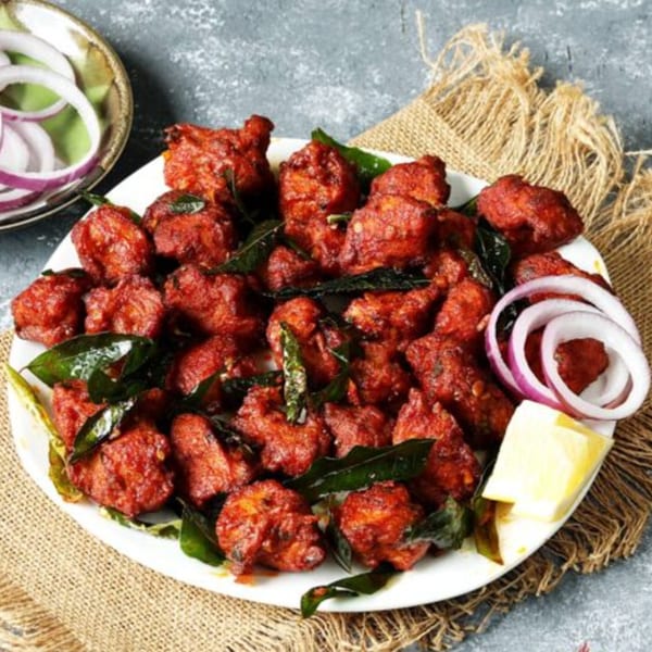 Red fried chicken pieces with curry leaves, onion rings, and lemon wedges on a white plate.