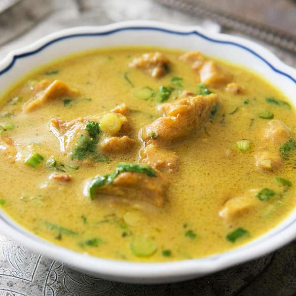 A close-up of a bowl of yellow curry with chicken and green herbs.