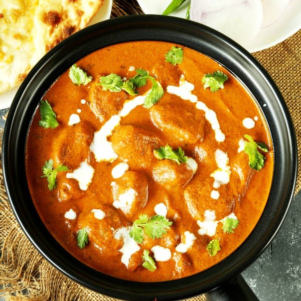 Overhead view of a black pan filled with butter chicken, garnished with cream and cilantro, alongside naan.