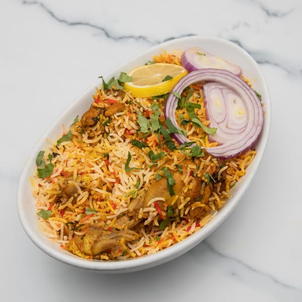 Mintt Indian Cuisine's chicken biryani, garnished with red onion, lemon, and cilantro, in a white bowl.