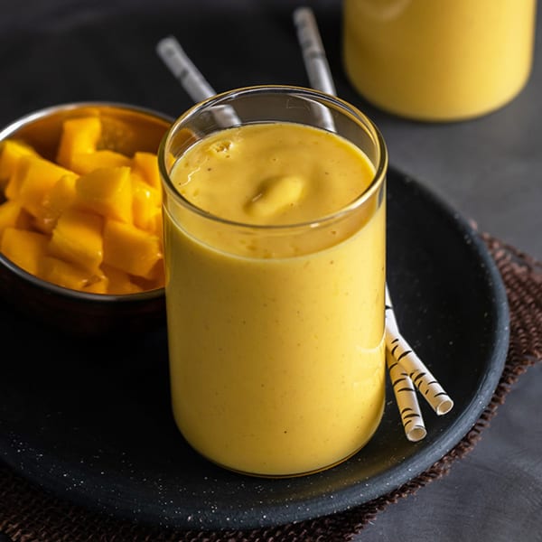 A glass of yellow mango lassi with a bowl of mango cubes and straws on a dark tray.