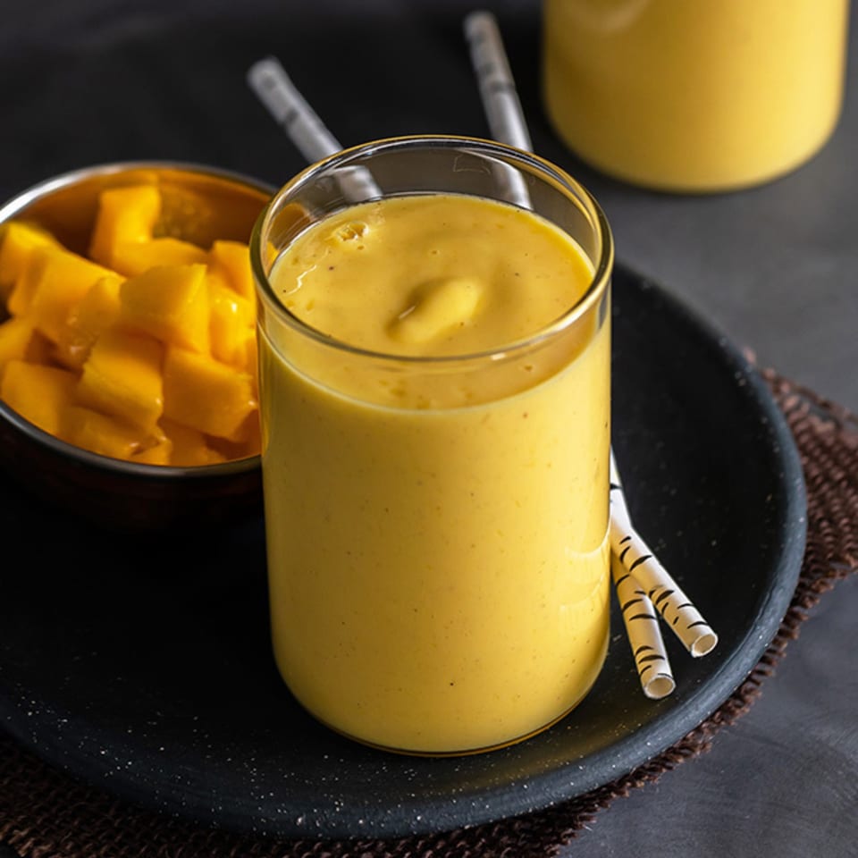 A glass of yellow mango lassi with a bowl of mango cubes and straws on a dark tray.