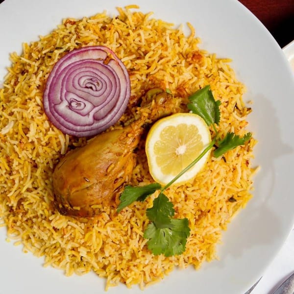 Chicken biryani with a drumstick, red onion slice, lemon wedge, and cilantro on a white plate.