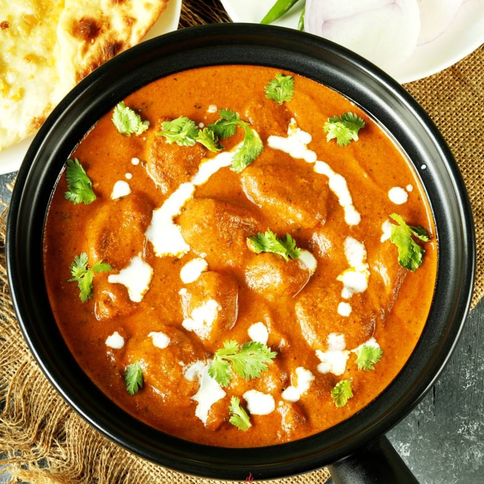 Overhead view of butter chicken in a black bowl, garnished with cream and cilantro, with naan bread.