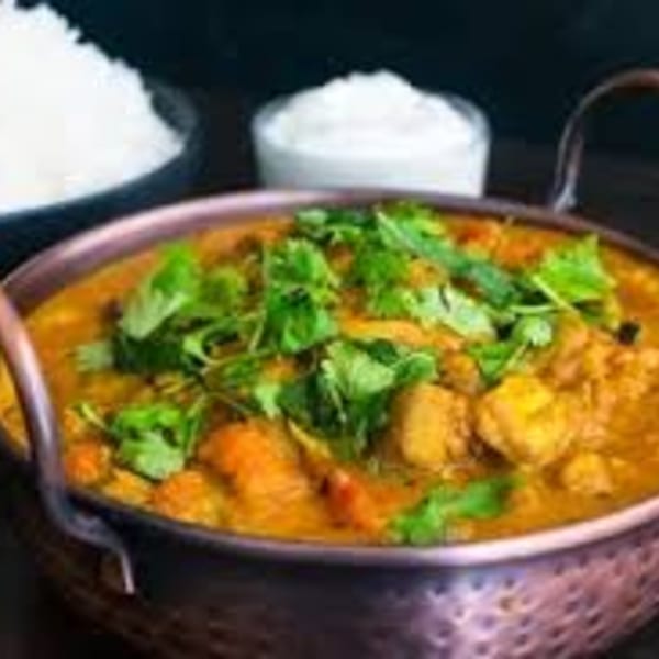 A copper pot filled with chicken curry, garnished with cilantro, served with rice and raita.
