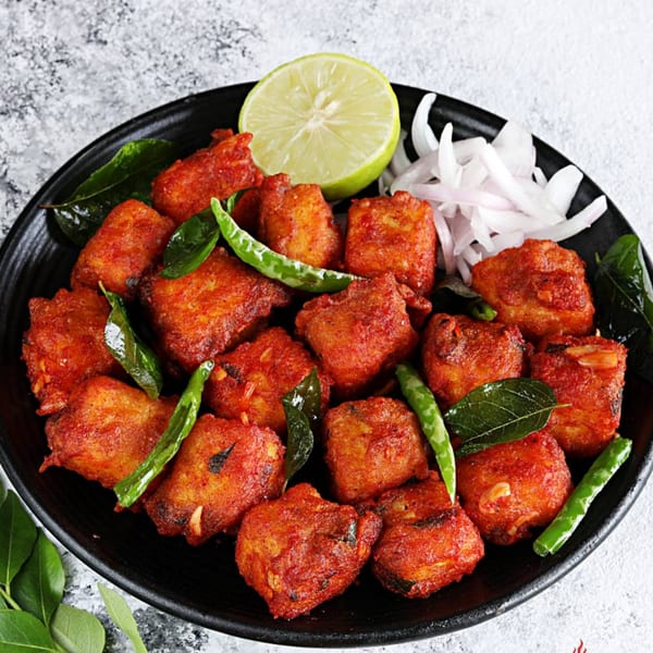 Spicy red fried chicken pieces garnished with green chilies, curry leaves, onions, and a lime wedge.