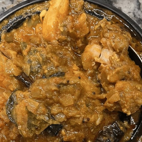 Close-up of a rich, golden-brown curry with chicken pieces, onions, and dark spices in a black bowl.