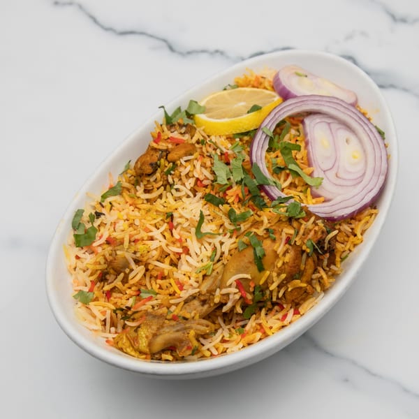 Mintt Indian Cuisine's chicken biryani, garnished with red onion, lemon, and cilantro in a white bowl.