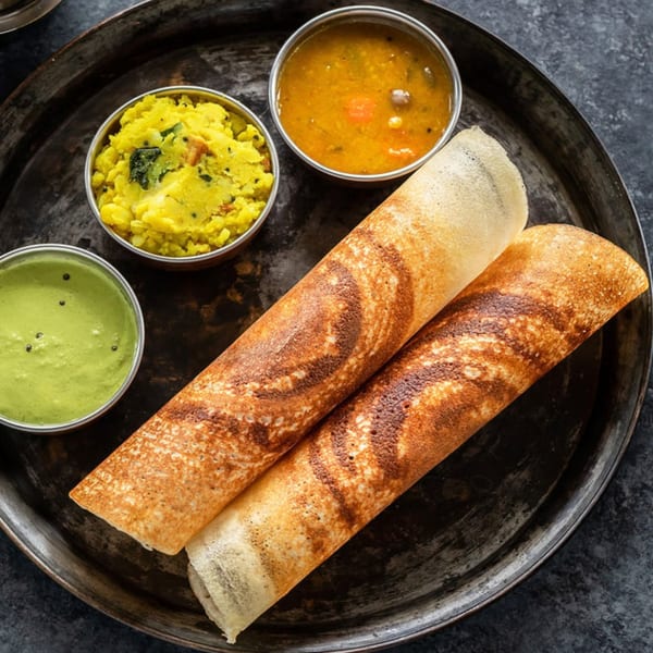 Two golden-brown dosas with bowls of green chutney, yellow potato masala, and sambar on a dark metal tray.