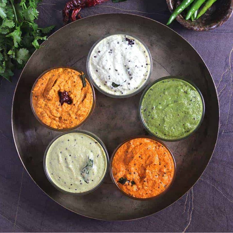 Five bowls of colorful chutneys, including green, white, and orange varieties, on a dark metal tray.