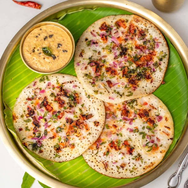 Three uttapam, topped with onions and herbs, served with chutney on a banana leaf.