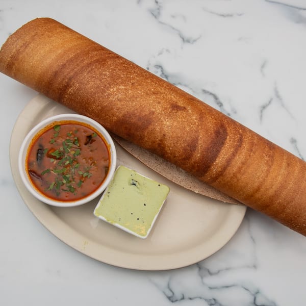 Long, rolled dosa with two small bowls of red and green chutneys on a white plate.