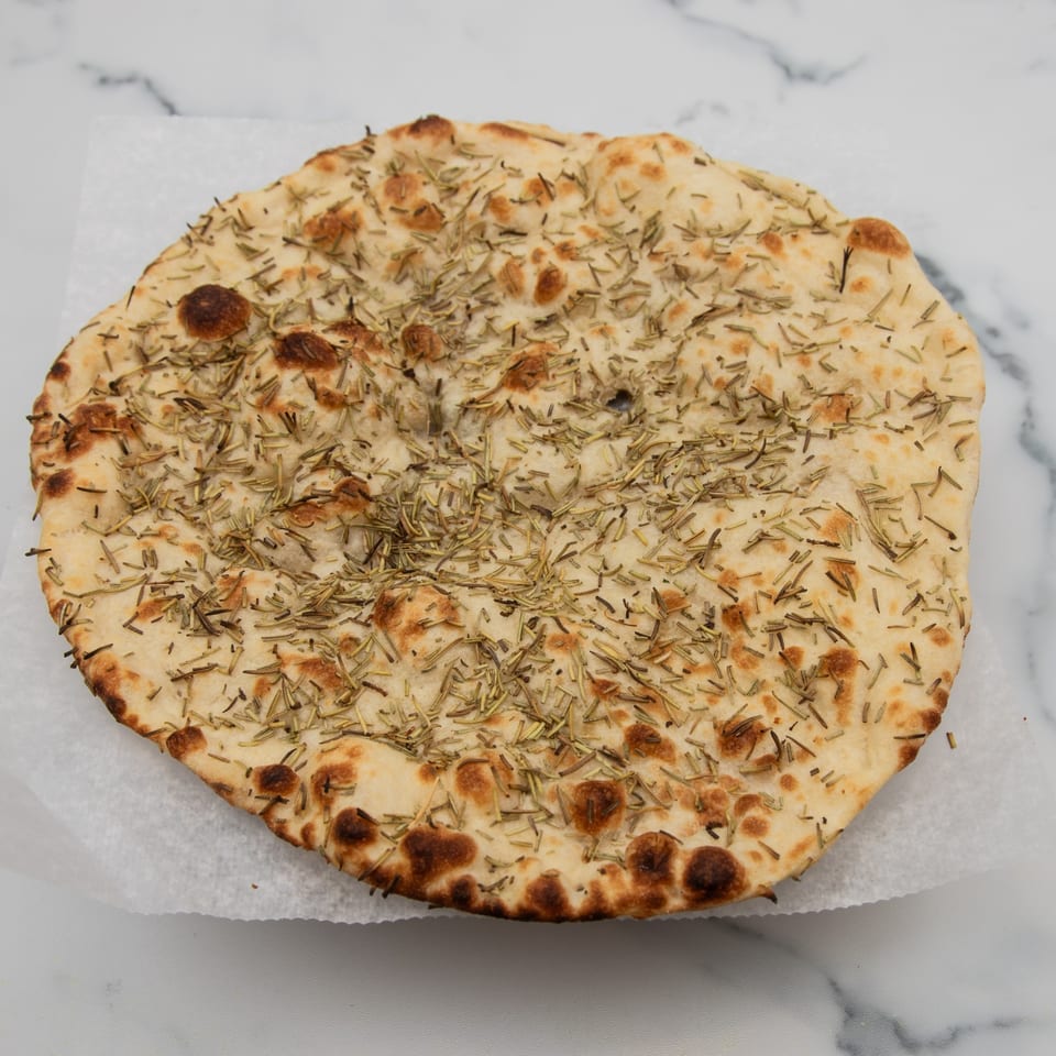 Round Indian flatbread, golden brown with dark spots, covered in dried rosemary on white parchment paper.