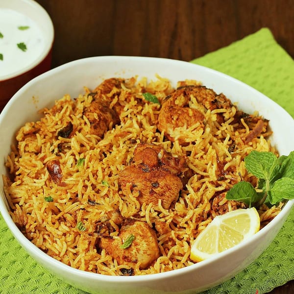 Chicken biryani in a white bowl with a lemon wedge, mint, and a side of raita.