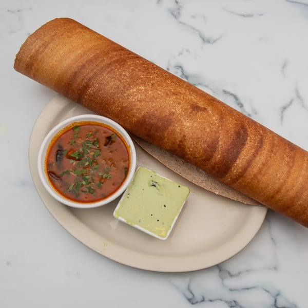 A large, rolled-up brown dosa with two small bowls of chutney and sambar on a white plate.