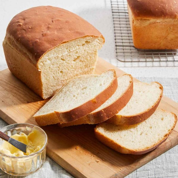 A freshly baked loaf of white bread, partially sliced, with a small bowl of butter and knife.