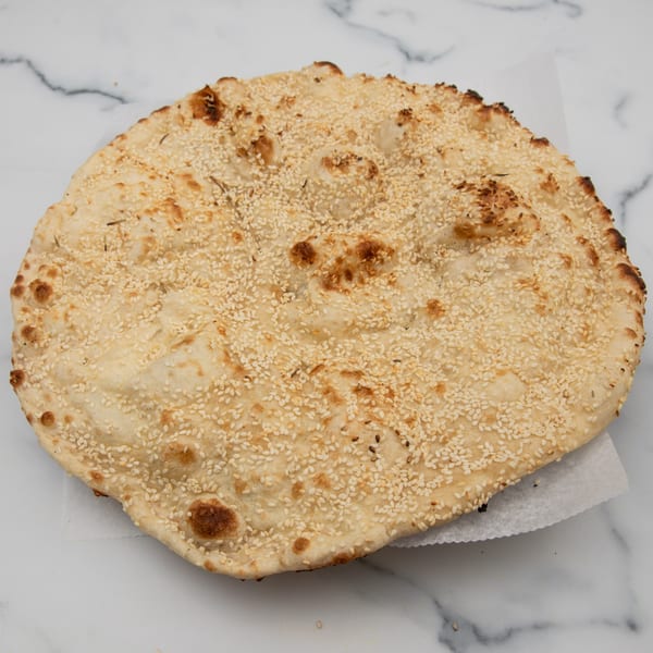 Round, golden-brown Indian naan bread, generously topped with sesame seeds, on a white marble surface.