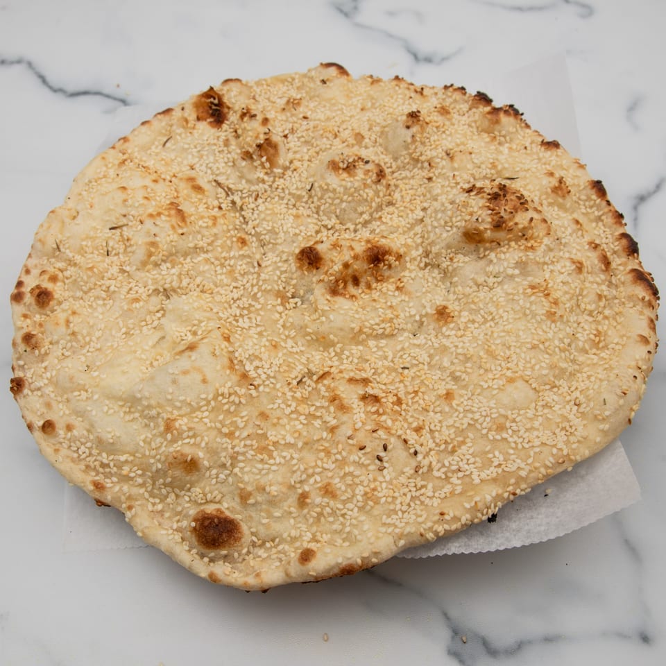 Round, golden-brown Indian naan bread, generously topped with sesame seeds, on a white marble surface.