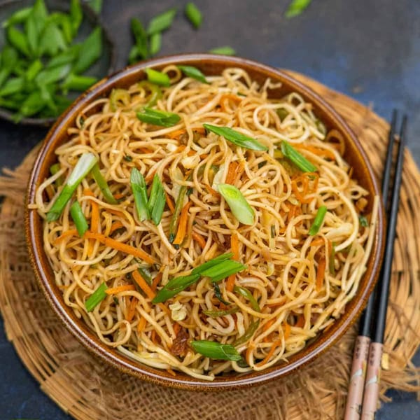 Close-up of a bowl of vegetable noodles, garnished with chopped green onions, on a woven placemat.
