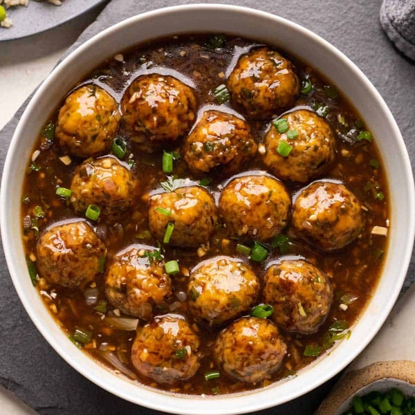 A white bowl filled with Manchurian vegetable balls in a rich brown gravy, garnished with green onions.