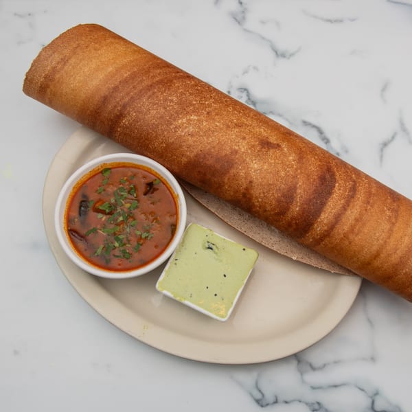 A large, crispy brown dosa roll served with a bowl of red sambar and green chutney.