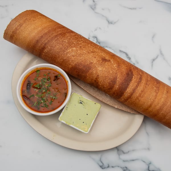 A long, rolled-up brown dosa served with a bowl of red curry and a small dish of green chutney.