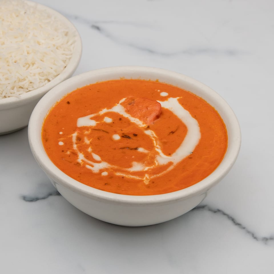A bowl of Mintt Indian Cuisine's orange butter chicken with cream swirls, next to white rice.