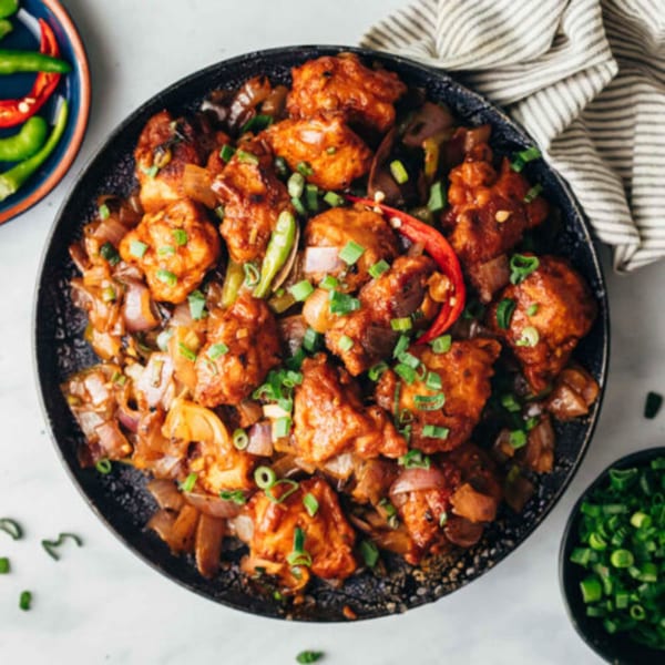 Spicy chicken Manchurian with onions, green onions, and red chili peppers in a black bowl.