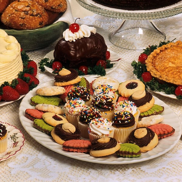 A white platter filled with assorted colorful cookies and cupcakes with sprinkles, surrounded by other desserts.