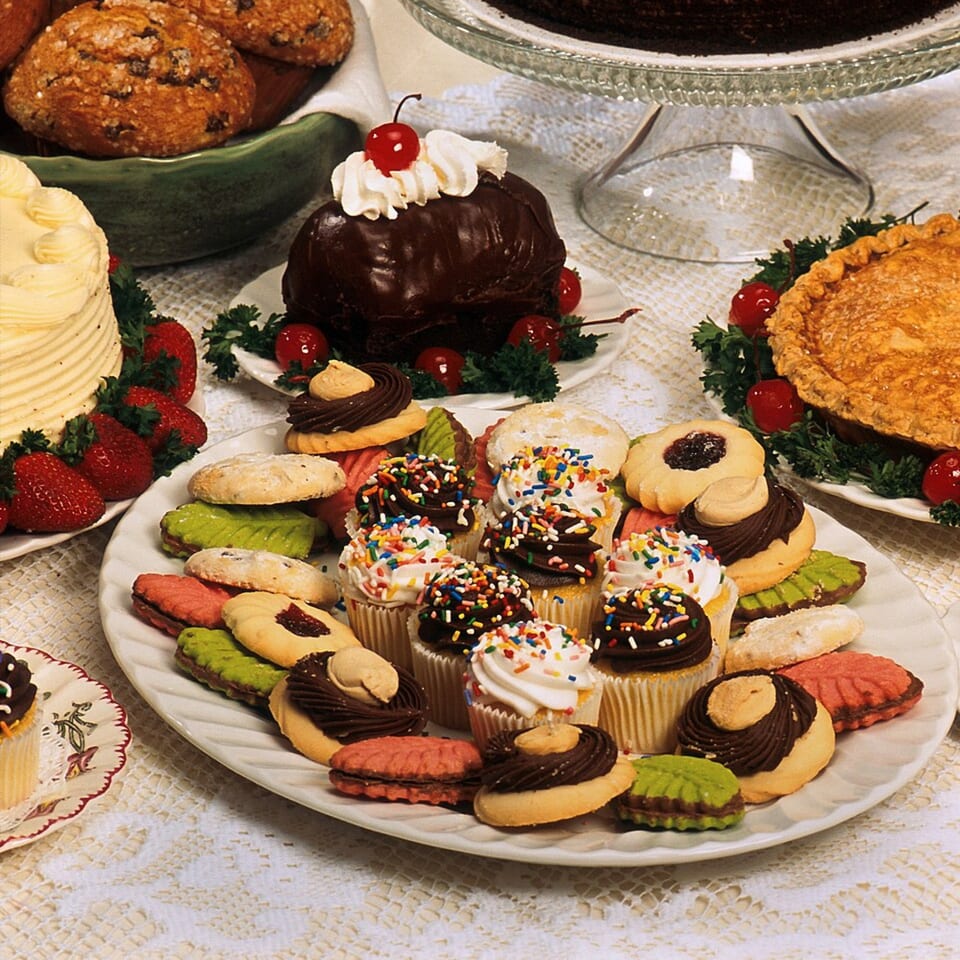 A white platter filled with assorted colorful cookies and cupcakes with sprinkles, surrounded by other desserts.