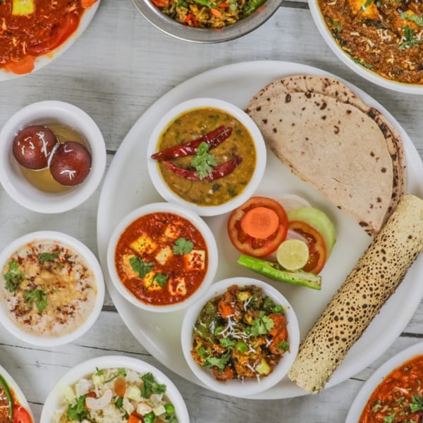 A white platter with various Indian dishes, including dal, paneer, vegetables, roti, and papad.