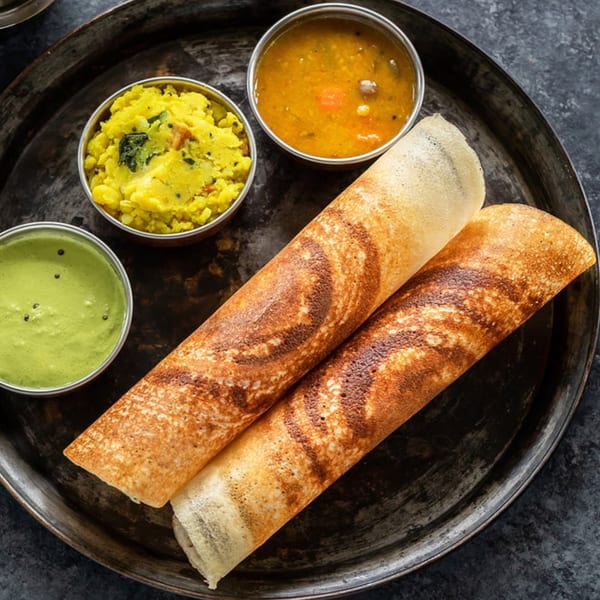 Two golden-brown dosas with bowls of green chutney, yellow potato curry, and sambar on a metal tray.