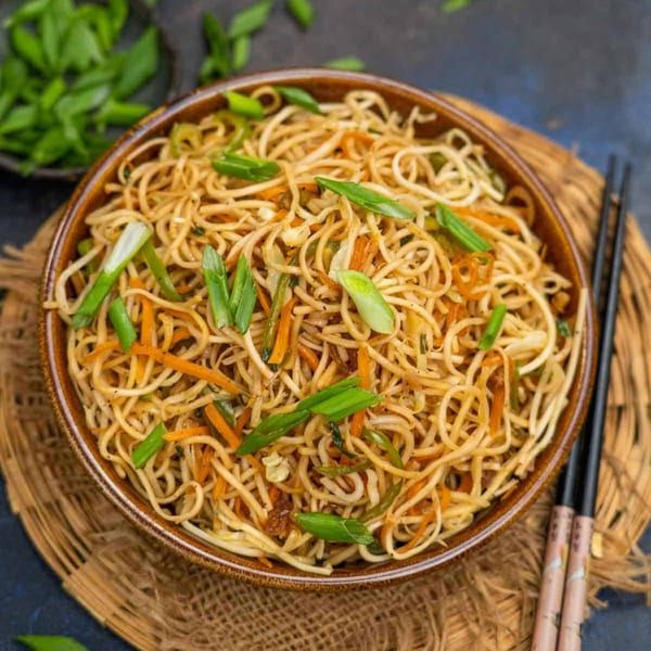 Close-up of a bowl of vegetable lo mein noodles, garnished with green onions, on a woven placemat.
